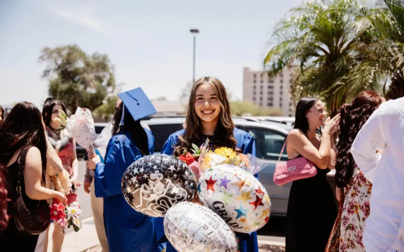 Génesis, vestida con un traje de graduación azul y sosteniendo globos y un ramo de flores, sonríe alegremente mientras celebra su graduación al aire libre con amigos y familiares.