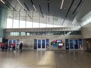 Vestíbulo del Centro de Ciencias de Arizona con grandes ventanas, paredes de hormigón y expositores a lo largo de la pared trasera; un grupo de visitantes se encuentra a la izquierda, cerca de una exposición.