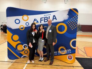 Tres personas posan delante de un gran telón azul, blanco y naranja con el logotipo de la FBLA de Arizona en un evento celebrado en un gimnasio cubierto. La persona del centro sostiene una medalla con una cinta roja. Las tres personas visten traje de negocios y llevan tarjetas identificativas del evento. En el fondo, a la izquierda del gimnasio, se puede ver a otros asistentes.