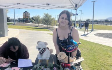 Una mujer sonríe junto a una mesa en la que se exponen accesorios para perros, bajo una glorieta en un parque.
